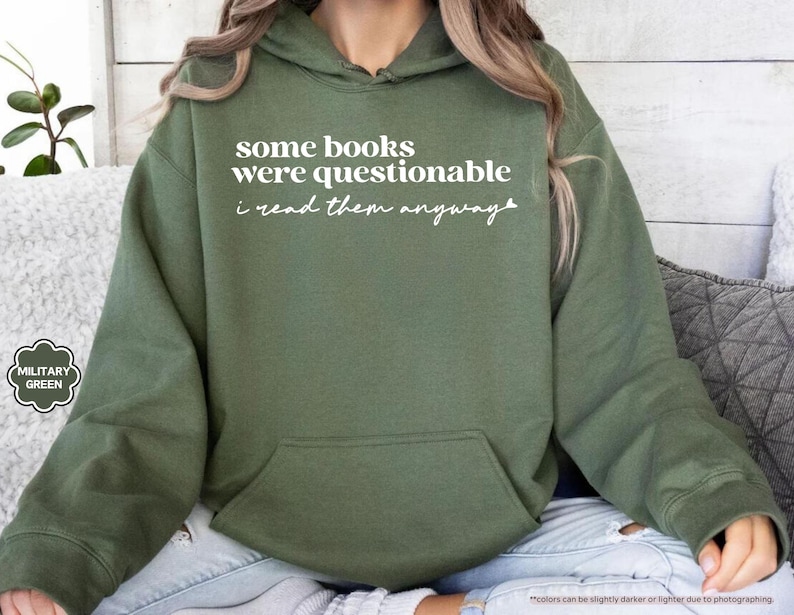 May include: A military green hooded sweatshirt featuring the white printed text some books were questionable i read them anyway. The garment includes a front kangaroo pocket, long sleeves with ribbed cuffs, and a drawstring hood in a matching color.
