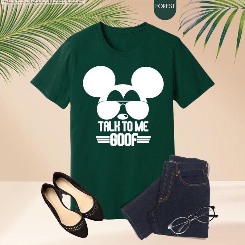 May include: Forest green t-shirt with a white Mickey Mouse graphic wearing sunglasses and the text "TALK TO ME GOOF". The image also shows black flats, dark wash jeans, and a pair of eyeglasses.