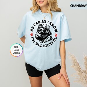 May include: Light blue t-shirt with a graphic of a gorilla wearing a cap, surrounded by the text "AS FAR AS I KNOW I'M DELIGHTFUL". The design is in black and white. The shirt has short sleeves.