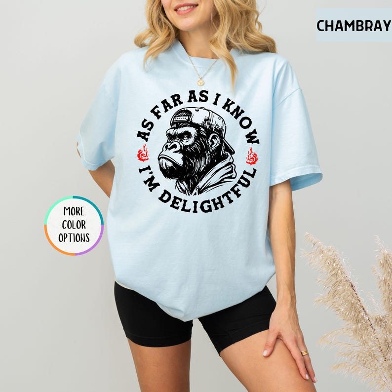 May include: Light blue t-shirt with a graphic of a gorilla wearing a cap, surrounded by the text "AS FAR AS I KNOW I'M DELIGHTFUL". The design is in black and white. The shirt has short sleeves.