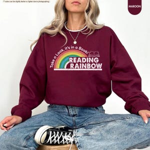 May include: Maroon sweatshirt with the text "Take a Look, it's in a Book! READING RAINBOW" and a rainbow graphic. The sweatshirt is a solid maroon color. The text is white with a rainbow graphic. The sweatshirt is a crew neck style.