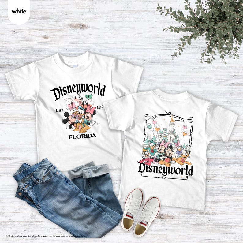 May include: Two white t-shirts with Disneyworld graphics. One shirt features Mickey Mouse and friends with "Disneyworld Florida Est 197" text. The other shirt has a castle and characters with "Disneyworld 1971" text. Blue jeans and white sneakers are also in the image.