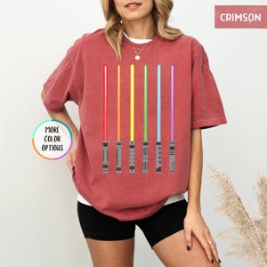 K&ouml;nnte beinhalten: Karmesinrotes T-Shirt mit einem Design aus sechs farbenfrohen Lichtschwertern in Rot, Orange, Gelb, Gr&uuml;n, Blau und Lila. Das Shirt hat eine lockere Passform und kurze &Auml;rmel. Die Worte "MORE COLOR OPTIONS" befinden sich in einem Kreis auf der linken Seite.