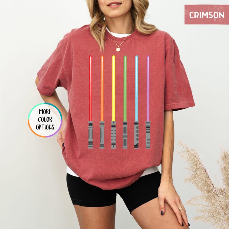 K&ouml;nnte beinhalten: Karmesinrotes T-Shirt mit einem Design aus sechs farbenfrohen Lichtschwertern in Rot, Orange, Gelb, Gr&uuml;n, Blau und Lila. Das Shirt hat eine lockere Passform und kurze &Auml;rmel. Die Worte "MORE COLOR OPTIONS" befinden sich in einem Kreis auf der linken Seite.
