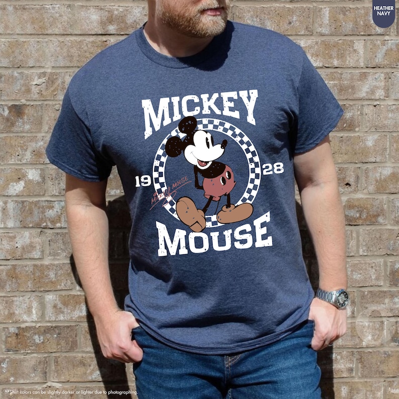 May include: Heather navy blue t-shirt featuring a graphic of Mickey Mouse. The design includes the text "MICKEY MOUSE" and the years "19" and "28". The cartoon character is in a circular frame with a checkered pattern.