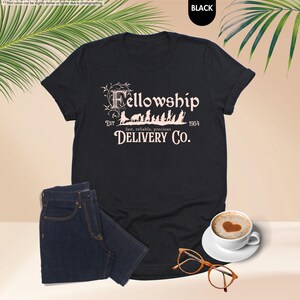 May include: Black t-shirt with the text "Fellowship Delivery Co." in a stylized font. The design includes silhouettes of figures and animals. A pair of dark blue jeans, a cup of coffee with a heart design, and glasses are also visible.