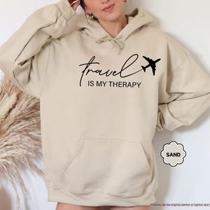 Pu&ograve; includere: Felpa con cappuccio color sabbia con tasca frontale e cappuccio con coulisse. Il testo nero recita "travel IS MY THERAPY" con una piccola grafica di aeroplano. La felpa con cappuccio &egrave; di un colore neutro, adatta all'abbigliamento casual.