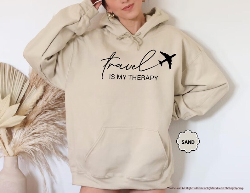 Pu&ograve; includere: Felpa con cappuccio color sabbia con tasca frontale e cappuccio con coulisse. Il testo nero recita "travel IS MY THERAPY" con una piccola grafica di aeroplano. La felpa con cappuccio &egrave; di un colore neutro, adatta all'abbigliamento casual.