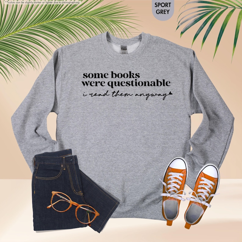 May include: A sport grey crewneck sweatshirt featuring the black text some books were questionable i read them anyway, paired with dark blue denim shorts, orange canvas sneakers with white laces, and brown-rimmed eyeglasses.