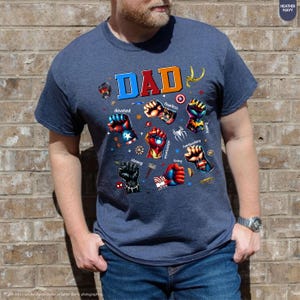 May include: Heather navy blue t-shirt with the word "DAD" in colorful letters and various superhero-themed fists. The shirt also features words like "devoted," "fearless," and "strong."