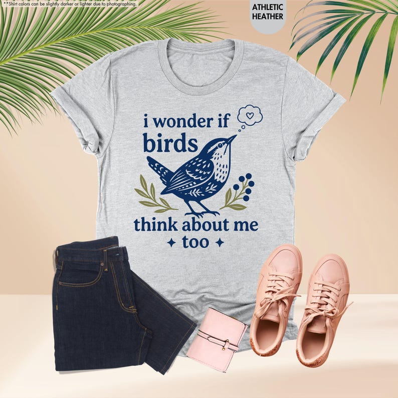 May include: An athletic heather gray short-sleeve t-shirt featuring a dark blue graphic of a bird with the text "i wonder if birds think about me too" and a small thought bubble with a heart, accompanied by dark blue denim jeans and pink sneakers.