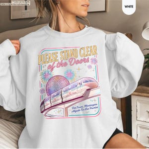 May include: White sweatshirt with a colorful graphic featuring a monorail, a geodesic sphere, and the text "PLEASE STAND CLEAR of the Doors." The design includes fireworks and the Spanish phrase "Por Favor, Mant&eacute;ngase Alejado De Las Puertas."