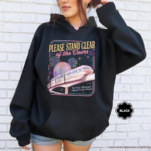 May include: Black hooded sweatshirt with a graphic featuring a monorail, fireworks, and the text "PLEASE STAND CLEAR of the Doors." The design includes a colorful illustration of a futuristic train and a large sphere. The word "BLACK" is also visible.