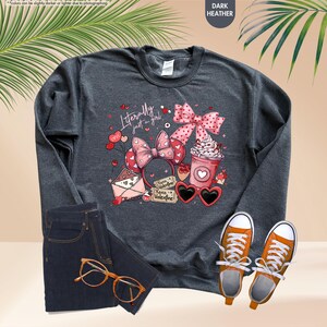 May include: Dark grey sweatshirt featuring the text "Literally just a girl" with a pink bow, heart-shaped sunglasses, a coffee cup, and a Minnie Mouse ear headband. The image also shows jeans and orange trainers.