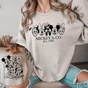 May include: Sand-colored sweatshirt featuring a black graphic of Mickey Mouse, Minnie Mouse, Donald Duck, and Daisy Duck. The text "MICKEY & CO Est. 1928" is printed below the characters. A smaller image of Mickey Mouse and friends is also visible.