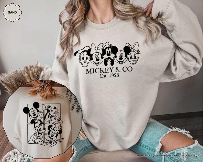 May include: Sand-colored sweatshirt featuring a black graphic of Mickey Mouse, Minnie Mouse, Donald Duck, and Daisy Duck. The text "MICKEY & CO Est. 1928" is printed below the characters. A smaller image of Mickey Mouse and friends is also visible.