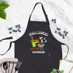 May include: Black apron with a cartoon image of Goofy grilling, the words "GRILL MASTER" and the name "ANDREW" in white. The apron has a neck strap and waist ties. The background includes blueberries and cooking utensils.
