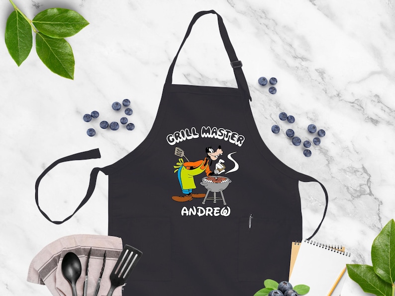 May include: Black apron with a cartoon image of Goofy grilling, the words "GRILL MASTER" and the name "ANDREW" in white. The apron has a neck strap and waist ties. The background includes blueberries and cooking utensils.