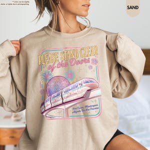 May include: Sand-colored sweatshirt featuring a colorful graphic with the text "PLEASE STAND CLEAR of the Doors". The design includes a monorail, a geodesic sphere, fireworks, and text in Spanish. A casual, comfortable top.