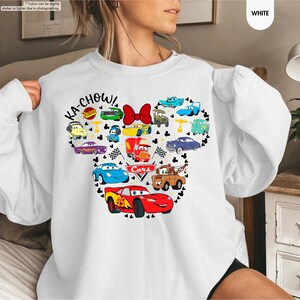 May include: White sweatshirt featuring a graphic of a Mickey Mouse head made up of Cars characters. The text "KA-CHOW!" and "Cars" are included in the graphic.