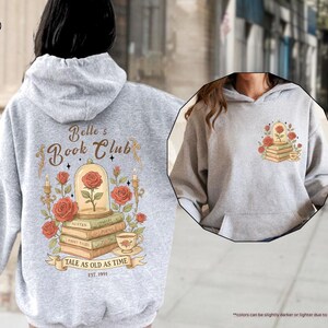 Puede incluir: Sudadera con capucha gris deportivo con un dise&ntilde;o que presenta una rosa en una c&uacute;pula de cristal, libros y el texto "Belle's Book Club". El dise&ntilde;o trasero incluye la frase "Tale as Old as Time" y el a&ntilde;o "EST. 1991".