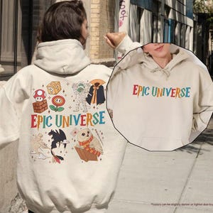 May include: A sand-colored hoodie with the text "EPIC UNIVERSE" in colorful letters. The back of the hoodie features various cartoon graphics, including a mushroom, a question mark block, and a mummy. The hoodie has a drawstring hood.