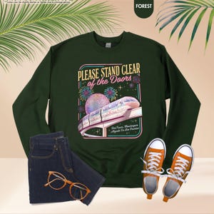 May include: Forest green sweatshirt with the text "PLEASE STAND CLEAR of the Doors" and a graphic of a monorail and fireworks. The image also includes a pair of blue jeans, orange sneakers, and glasses.