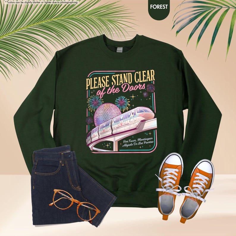 May include: Forest green sweatshirt with the text "PLEASE STAND CLEAR of the Doors" and a graphic of a monorail and fireworks. The image also includes a pair of blue jeans, orange sneakers, and glasses.