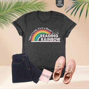 May include: Dark grey heather t-shirt with the text "Take a Look, it's in a Book! Reading Rainbow" and a rainbow graphic. The image also shows a pair of blue jeans, a small pink wallet, and a pair of pink sneakers.