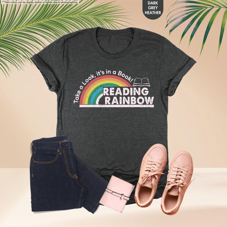 May include: Dark grey heather t-shirt with the text "Take a Look, it's in a Book! Reading Rainbow" and a rainbow graphic. The image also shows a pair of blue jeans, a small pink wallet, and a pair of pink sneakers.