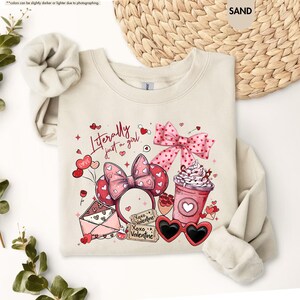 May include: A sand-coloured sweatshirt with a Valentine's Day design. The graphic includes a pink bow, heart-shaped sunglasses, a coffee cup, and the text "Literally just a girl." The design also features a pair of Minnie Mouse ears.