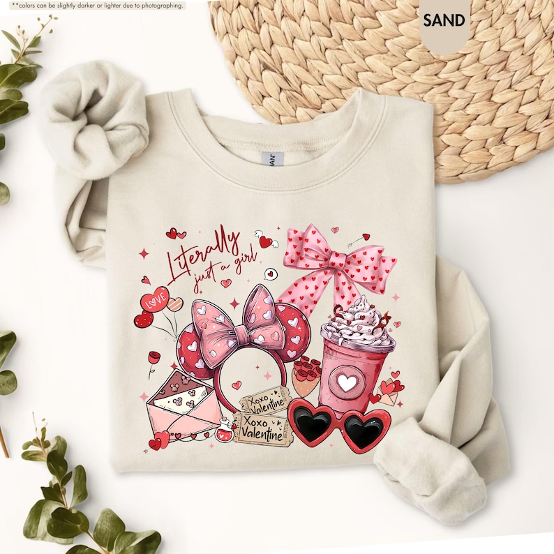 May include: A sand-coloured sweatshirt with a Valentine's Day design. The graphic includes a pink bow, heart-shaped sunglasses, a coffee cup, and the text "Literally just a girl." The design also features a pair of Minnie Mouse ears.