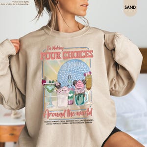 Pu&ograve; includere: Felpa color sabbia con la scritta "I'm Making Pour Choices" e "Around the World". La grafica presenta cocktail colorati e un design a sfera blu. La felpa elenca diversi paesi.