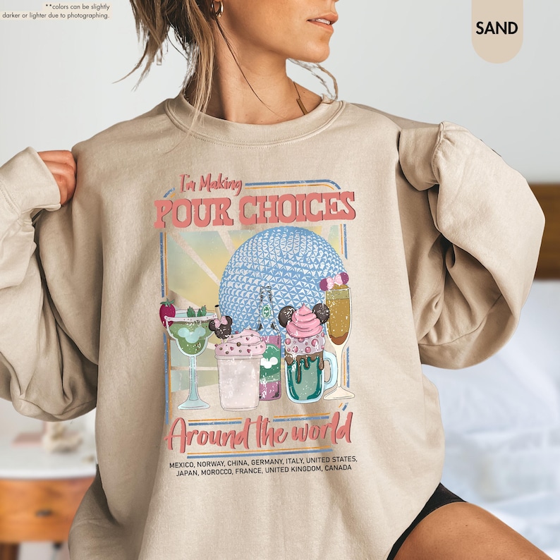 Pu&ograve; includere: Felpa color sabbia con la scritta "I'm Making Pour Choices" e "Around the World". La grafica presenta cocktail colorati e un design a sfera blu. La felpa elenca diversi paesi.