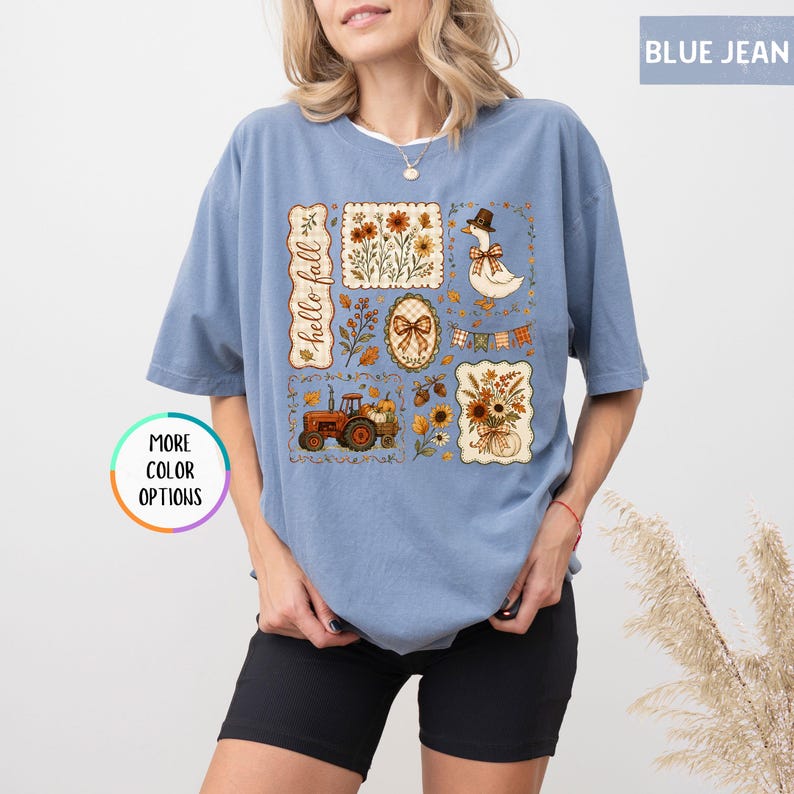 May include: A blue jean colored t-shirt featuring a graphic print with the text hello fall. The design includes a tractor, a goose wearing a hat, sunflowers, autumn leaves, and plaid bows arranged in a collage style on the front of the shirt.