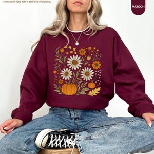 May include: Maroon sweatshirt with a floral design featuring white daisies, orange flowers, and pumpkins. The word "Maroon" is in the upper right corner. The sweatshirt is a long-sleeved crew neck.