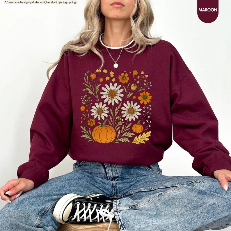 May include: Maroon sweatshirt with a floral design featuring white daisies, orange flowers, and pumpkins. The word "Maroon" is in the upper right corner. The sweatshirt is a long-sleeved crew neck.