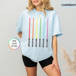 K&ouml;nnte beinhalten: Hellblaues T-Shirt mit einem Design aus sechs farbigen Lichtschwertern in Rot, Orange, Gelb, Gr&uuml;n, Blau und Lila. Auf dem Shirt steht "CHAMBRAY" oben rechts und "MORE COLOR OPTIONS" unten links.