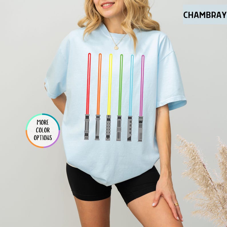 K&ouml;nnte beinhalten: Hellblaues T-Shirt mit einem Design aus sechs farbigen Lichtschwertern in Rot, Orange, Gelb, Gr&uuml;n, Blau und Lila. Auf dem Shirt steht "CHAMBRAY" oben rechts und "MORE COLOR OPTIONS" unten links.