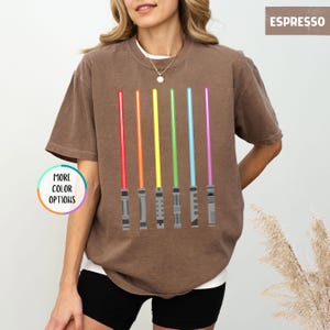K&ouml;nnte beinhalten: Espressofarbenes T-Shirt mit einem Design aus sechs farbigen Lichtschwertern in Rot, Orange, Gelb, Gr&uuml;n, Blau und Lila. Das Shirt hat eine lockere Passform und einen Rundhalsausschnitt. Die Worte "MORE COLOR OPTIONS" sind in einem Kreis links.