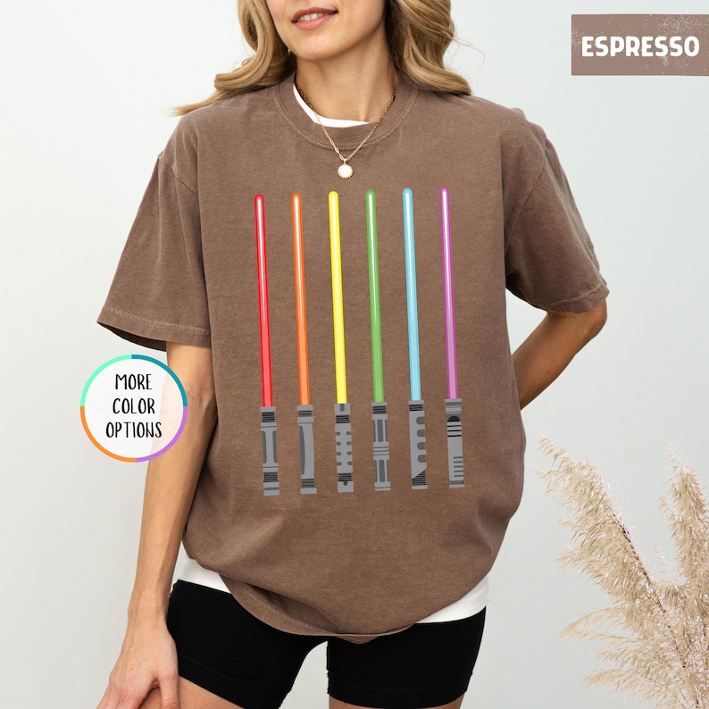 K&ouml;nnte beinhalten: Espressofarbenes T-Shirt mit einem Design aus sechs farbigen Lichtschwertern in Rot, Orange, Gelb, Gr&uuml;n, Blau und Lila. Das Shirt hat eine lockere Passform und einen Rundhalsausschnitt. Die Worte "MORE COLOR OPTIONS" sind in einem Kreis links.