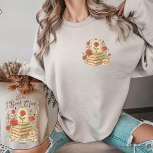 Puede incluir: Sudadera color arena con un dise&ntilde;o de una rosa en una c&uacute;pula de cristal sobre libros, rodeada de rosas y una taza de t&eacute;. El dise&ntilde;o incluye el texto "Belle's Book Club" y "Tale as Old as Time". La sudadera es de color neutro.