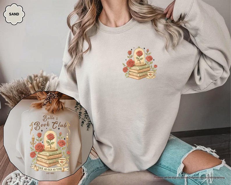 Puede incluir: Sudadera color arena con un dise&ntilde;o de una rosa en una c&uacute;pula de cristal sobre libros, rodeada de rosas y una taza de t&eacute;. El dise&ntilde;o incluye el texto "Belle's Book Club" y "Tale as Old as Time". La sudadera es de color neutro.