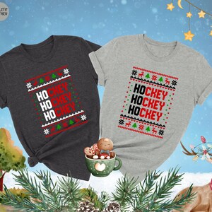 May include: Two Christmas themed t-shirts with the words "Hockey" repeated three times on each shirt. One shirt is dark heather grey and the other is light grey.