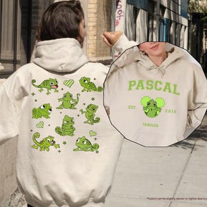 Op de afbeelding: Zandkleurige hoodie met groene kameleonafbeeldingen en het woord "PASCAL" in groen. De voorkant van de hoodie heeft een afbeelding van een kameleon met de tekst "EST. 2010 TANGLED".