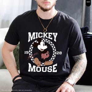May include: Black t-shirt featuring a graphic of Mickey Mouse. The design includes the word "MICKEY" above the character, and "MOUSE" below, with "1928" on either side of Mickey in a checkered circle. The shirt is a classic crew neck.