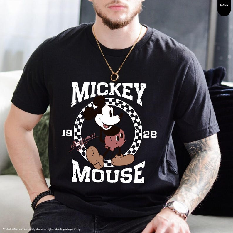 May include: Black t-shirt featuring a graphic of Mickey Mouse. The design includes the word "MICKEY" above the character, and "MOUSE" below, with "1928" on either side of Mickey in a checkered circle. The shirt is a classic crew neck.