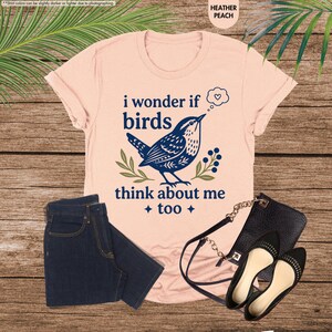 May include: A heather peach t-shirt featuring a dark blue graphic of a bird, green leaves, and the text "i wonder if birds think about me too" with a thought bubble containing a heart. Dark blue denim jeans and a black purse with black flats.