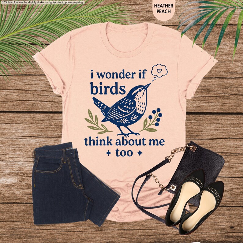 May include: A heather peach t-shirt featuring a dark blue graphic of a bird, green leaves, and the text "i wonder if birds think about me too" with a thought bubble containing a heart. Dark blue denim jeans and a black purse with black flats.