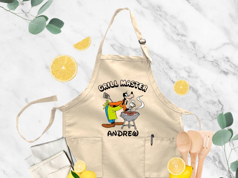 May include: A beige apron with a cartoon image of a dog grilling. The text "GRILL MASTER" is above the dog, and the name "ANDREW" is below. The apron has two pockets and a pen holder.